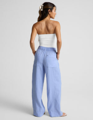 Woman wearing a white strapless top and blue linen pants against a plain background. 