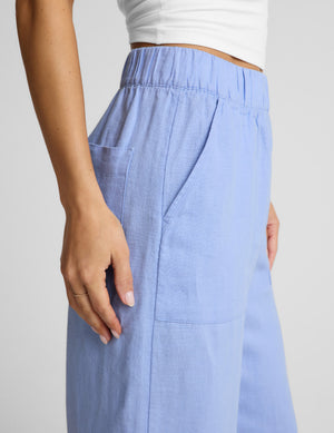 Woman wearing a white strapless top and blue linen pants against a plain background. 