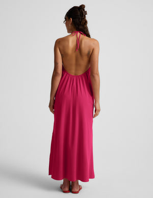 Woman wearing a red halter maxi dress with an open back against a light gray background. 