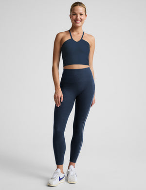 Woman wearing a navy blue cropped tank top and midi leggings with white contrast piping on the sides against a plain background. 
