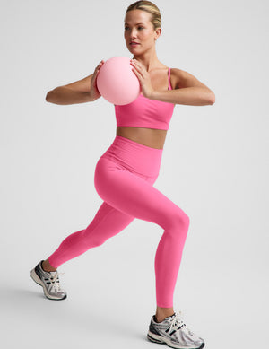 Woman in pink sports bra and leggings holding a pink exercise ball on a light gray background. 