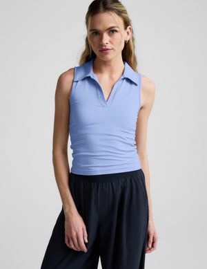 Woman wearing a light blue sleeveless polo shirt and black pants on a plain background. 