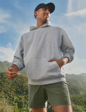 man wearing a gray hoodie and green shorts standing outdoors with mountains in the background. 