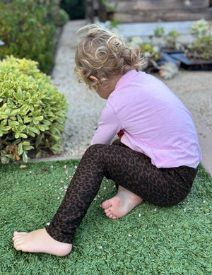 Toddler in pink sits on grass wearing Espresso Leopard SoftMark Toddler Legging featuring a dark brown leopard print pattern.