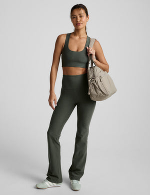 A woman wears olive Spacedye Practice High Waisted Bootcut Pant with a matching top and beige bag on a neutral background.