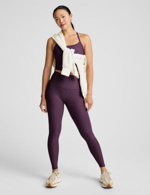 Woman wearing a purple cropped tank and leggings against a plain background. 