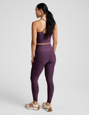 Woman wearing a purple cropped tank and leggings against a plain background. 