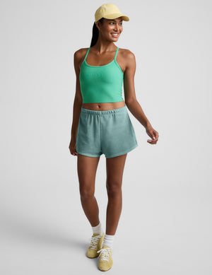 Woman wearing a green cropped tank and light blue shorts with a yellow cap on a plain background. 
