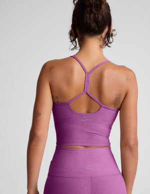 Woman wearing a purple cropped tank and leggings on a gray background. 
