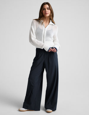 Woman wearing a white zip-up sweater and blue trousers against a plain background. 