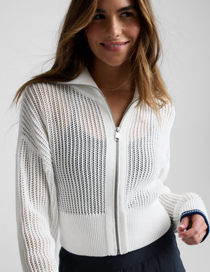 Woman wearing a white zip-up sweater and blue trousers against a plain background. 
