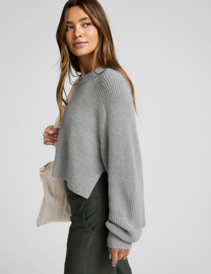 Woman wearing a gray sweater holding a beige bag against a white background. 
