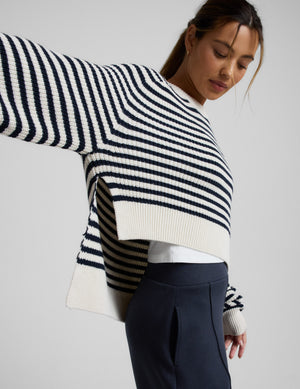Woman wearing a white and blue striped hi lo sweater and blue pants against a light gray background. 