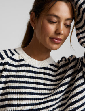 Woman wearing a white and blue striped hi lo sweater and blue pants against a light gray background. 