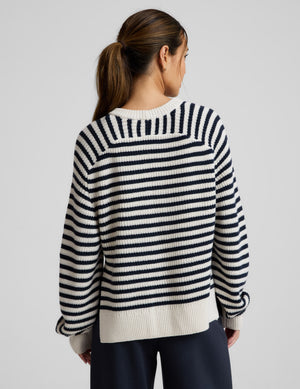 Woman wearing a white and blue striped hi lo sweater and blue pants against a light gray background. 